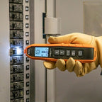 Person wearing a yellow glove holding an orange digital multimeter in front of electrical panels.