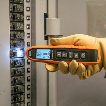 Person wearing a yellow glove holding an orange digital multimeter in front of electrical panels.