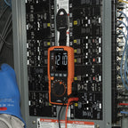 Multimeter on a panel with numbered circuits and a blue glove partially visible