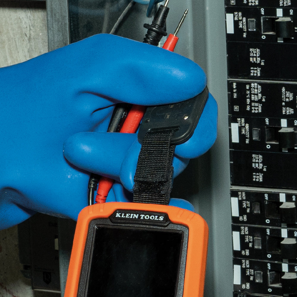 Person wearing blue gloves holding a Klein Tools multimeter in front of electrical panels.