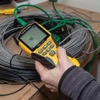 Hand holding a yellow and black device with a digital display in front of coiled cables.