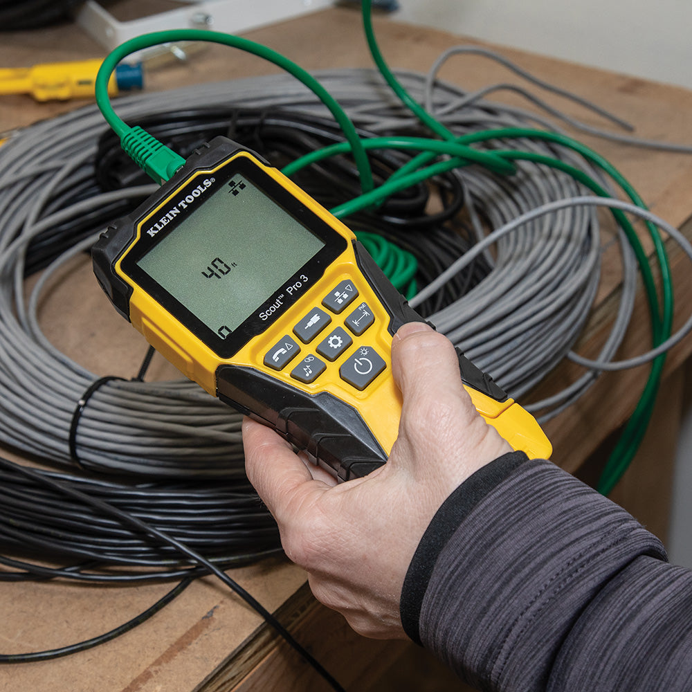 Hand holding a yellow and black device with a digital display in front of coiled cables.