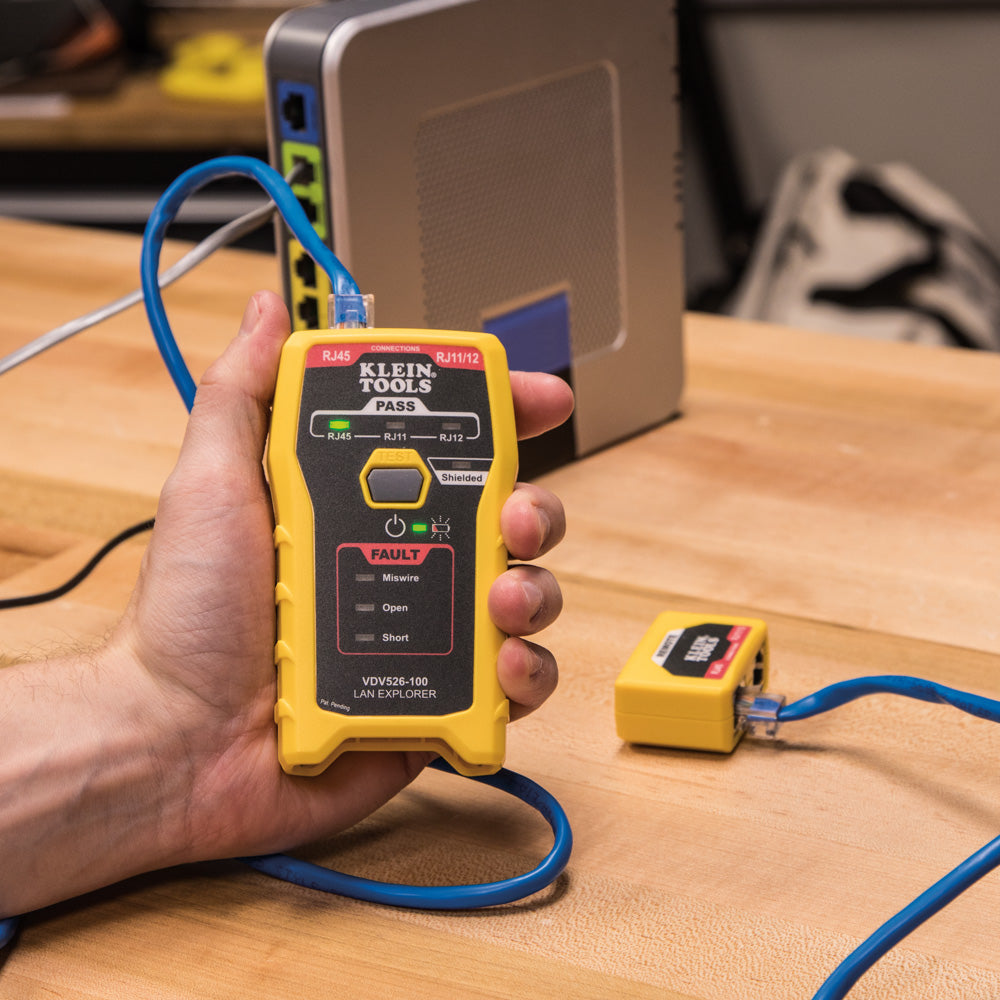 Hand holding a yellow diagnostic tool on a wooden surface with cables and equipment in the background.